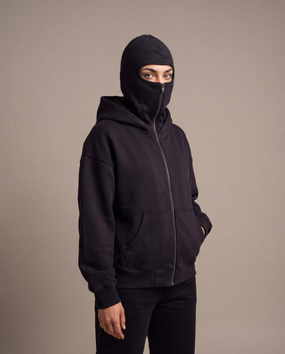 The Cover Hoodie in black, zipped up to just below the eyes, studio photo